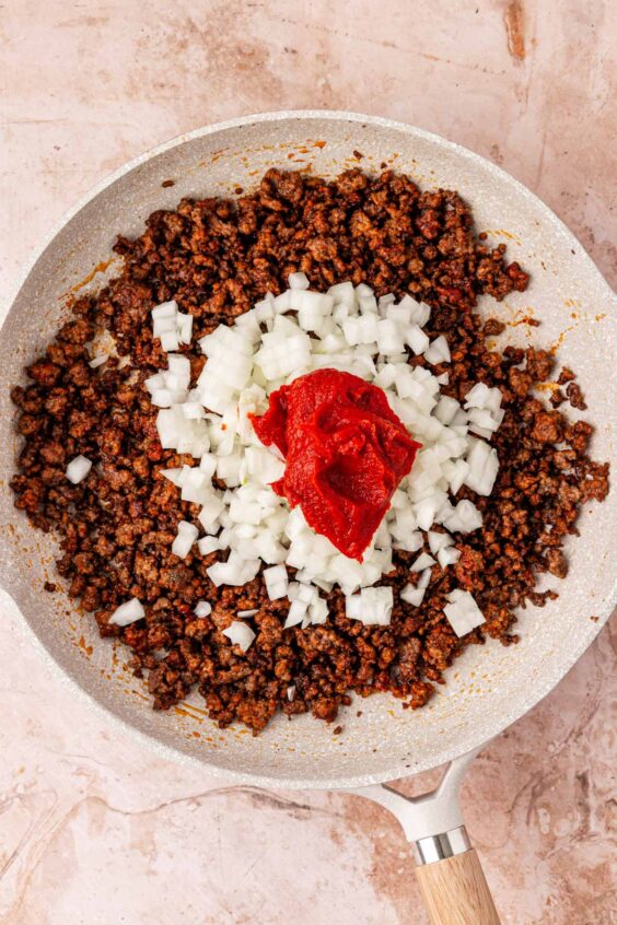 Onion and tomato paste being added to a skillet with ground beef.